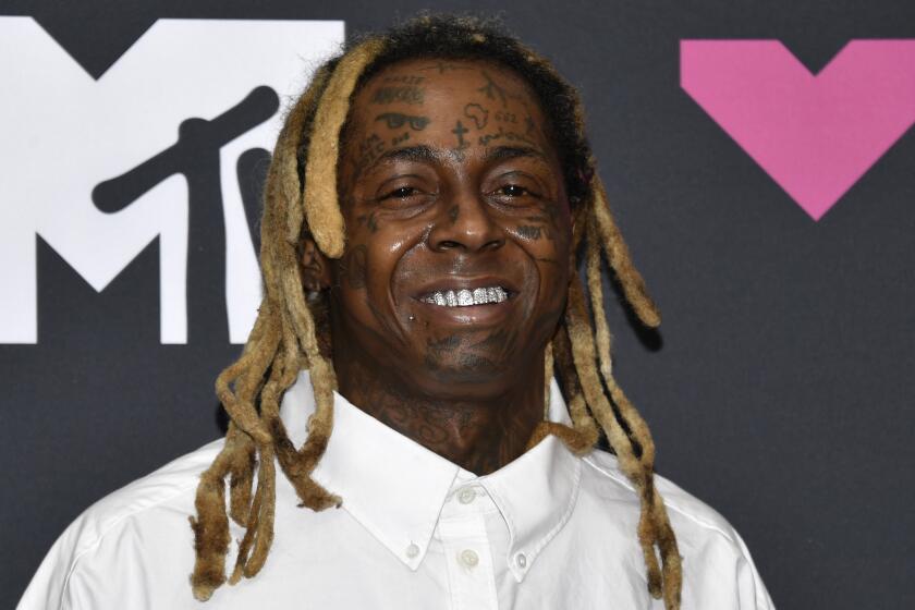 Lil Wayne in a white button down shirt smiling and posing in front of a black backdrop