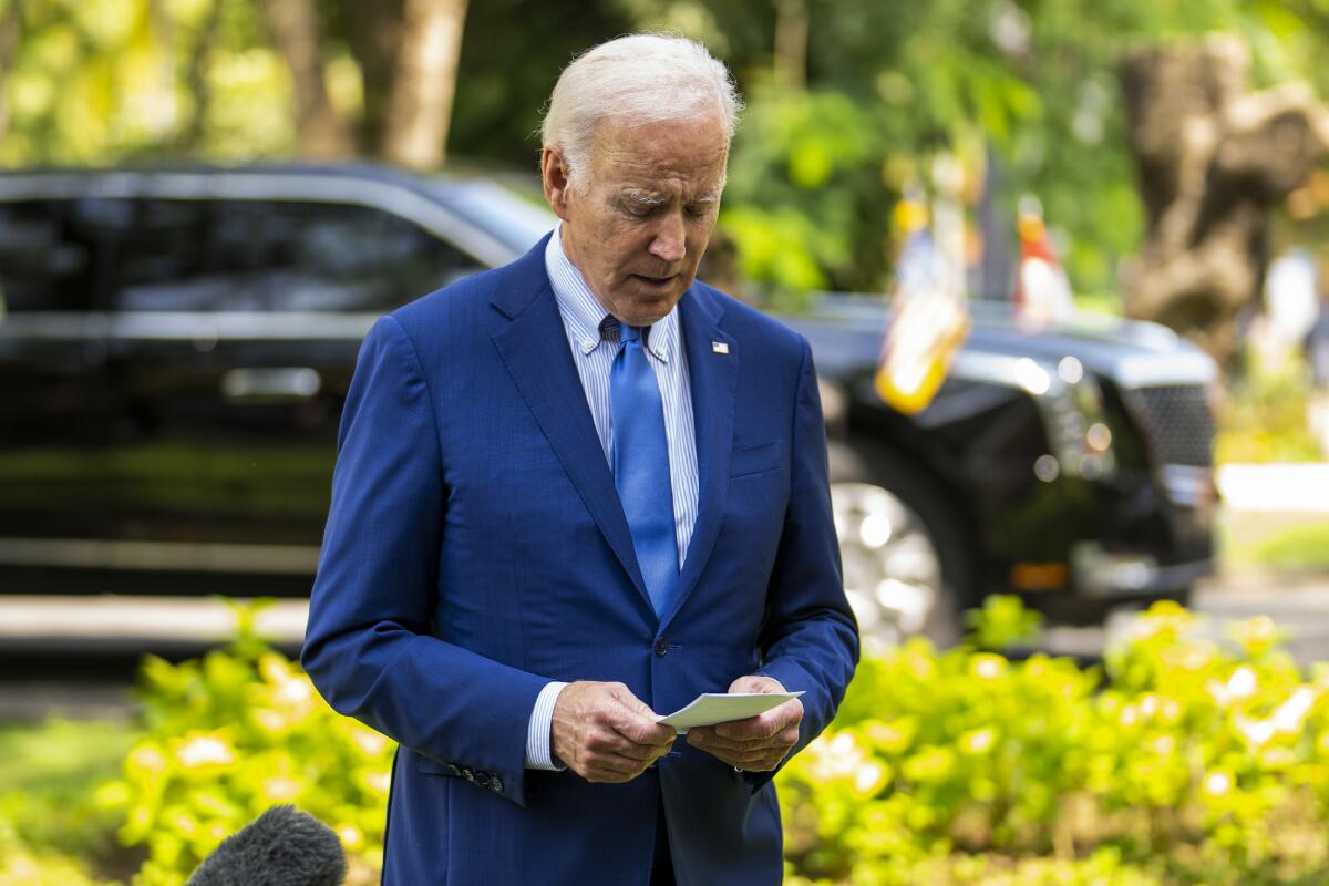 President Biden looking down at his notes as he speaks outside, standing in front of greenery and a large black vehicle
