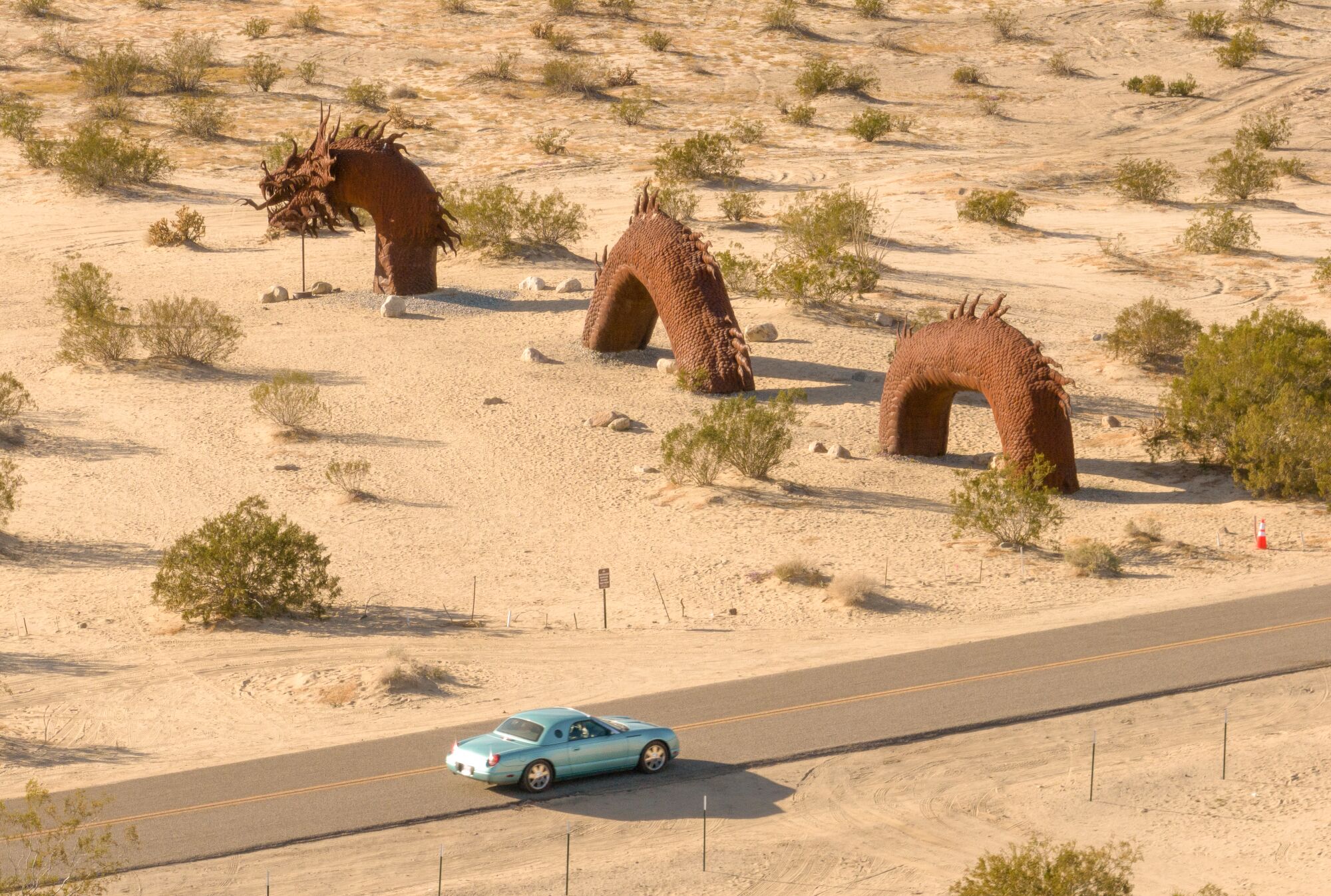An overhead image of a giant metallic sea serpent statue in the desert, as a blue car passes by on the road next to it.