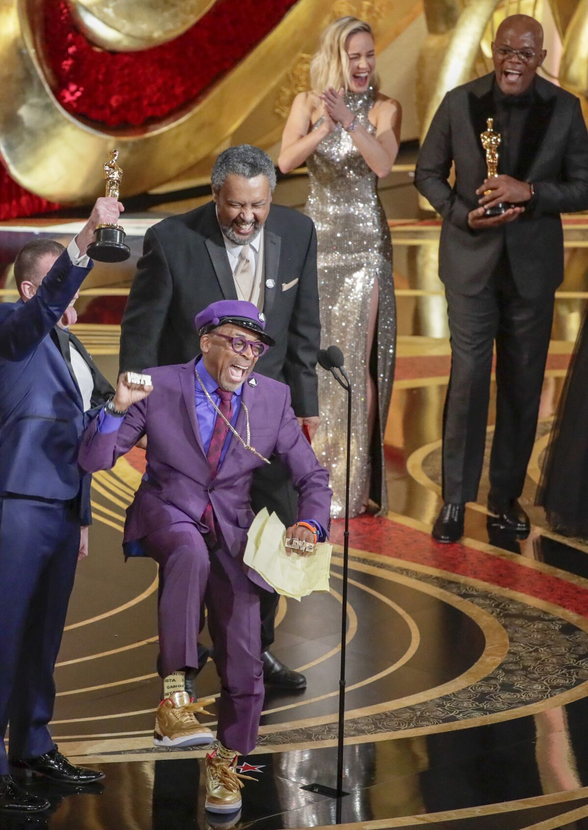 Spike Lee, in purple suit, reacts joyously after winning his first Oscar, presented by Brie Larson and Samuel L. Jackson, off to the right.