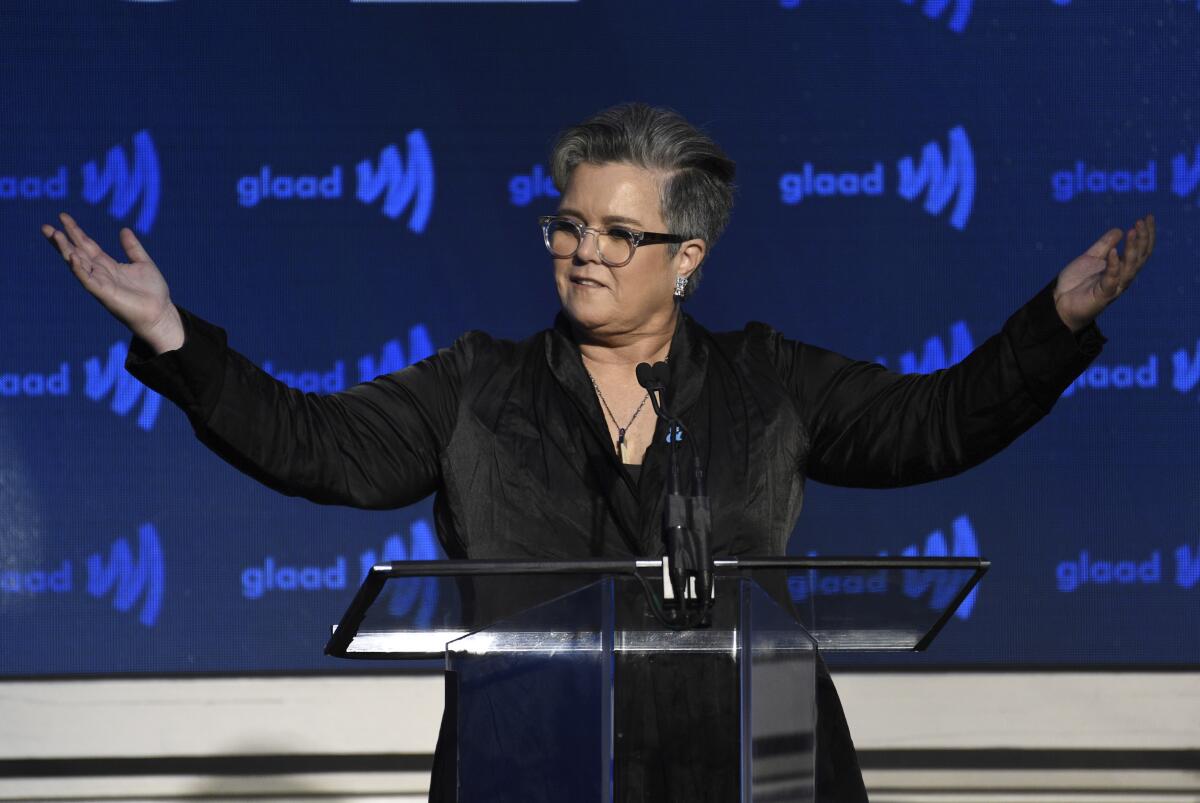 Rosie O'Donnell standing at a microphone wearing a black blazer with her arms outstretched and palms up