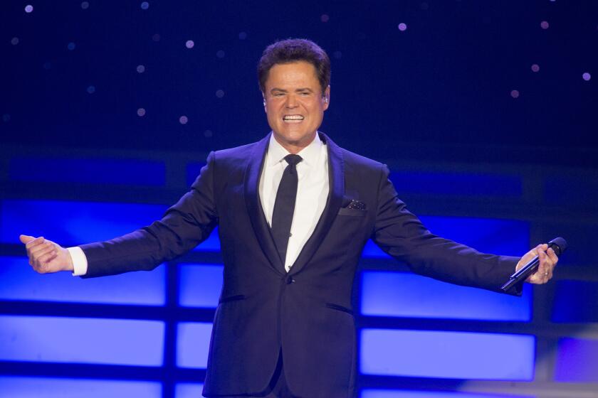 Donny Osmond on stage wearing a suit and tie smiling with his hands out to either side