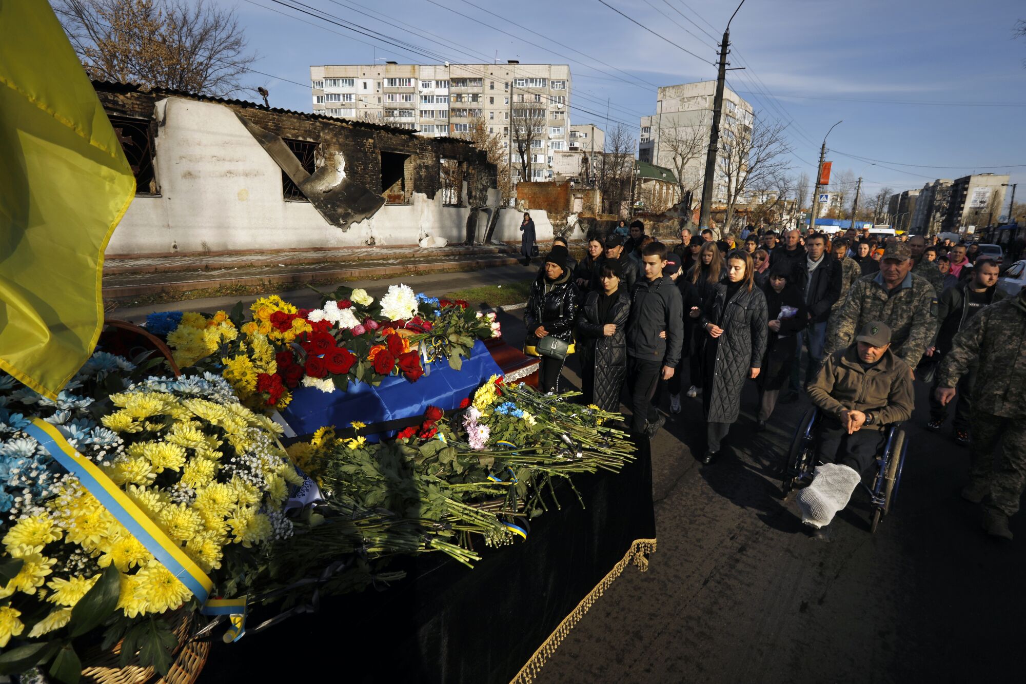 A group of people in dark clothes or military camouflage follow a flower-covered casket on a city street