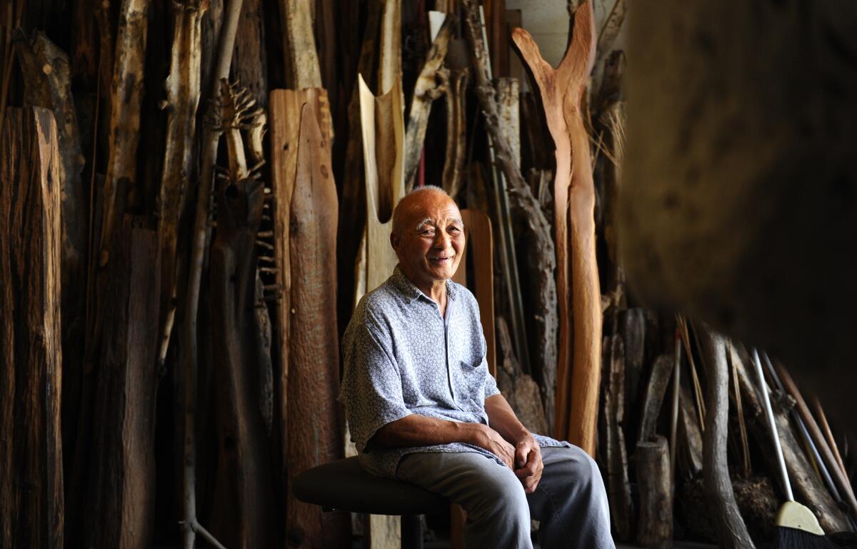 Kenzi Shiokova smiles as he sits before rows of tree trunks in various states of sculptural fabrication