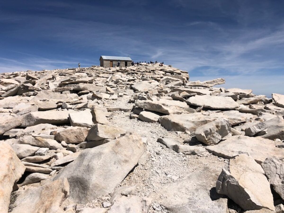 A view looking up a rocky hill. In the distance is a small dwelling surrounded by people.