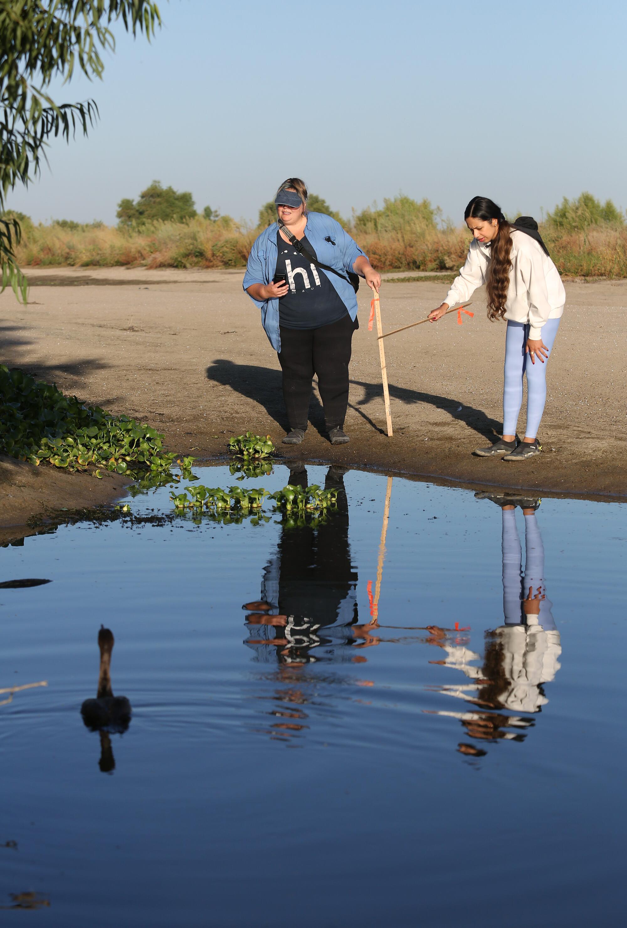 Two women look at a stagnant pool in a dry river bed.