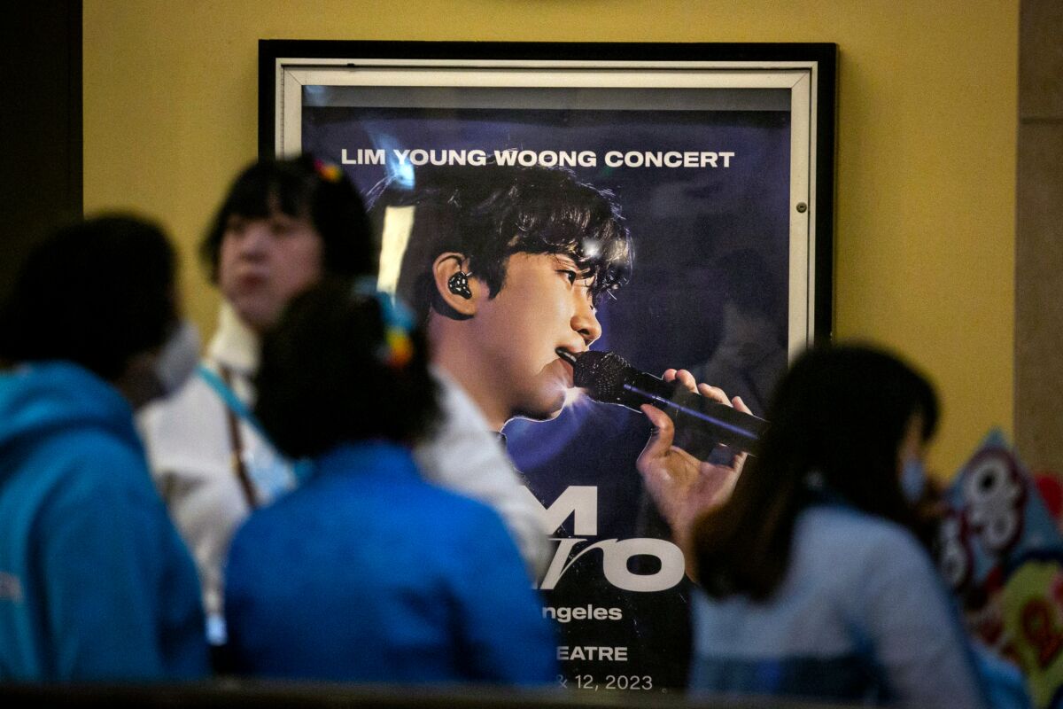Women in blue shirts gather near a poster of a young Korean man holding a microphone