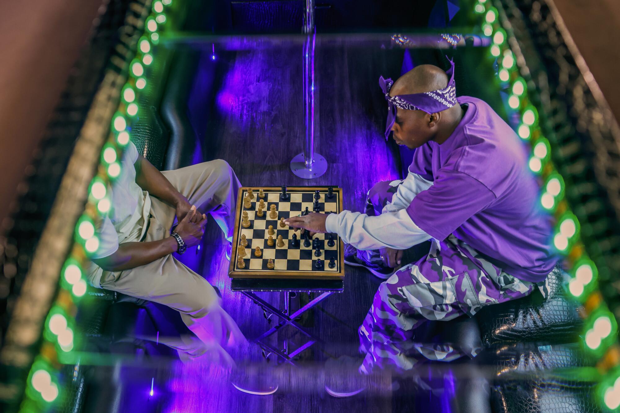 Vincent Hubbard dressed in a purple bandana and shirt plays chess in a party bus lit with purple lights. 