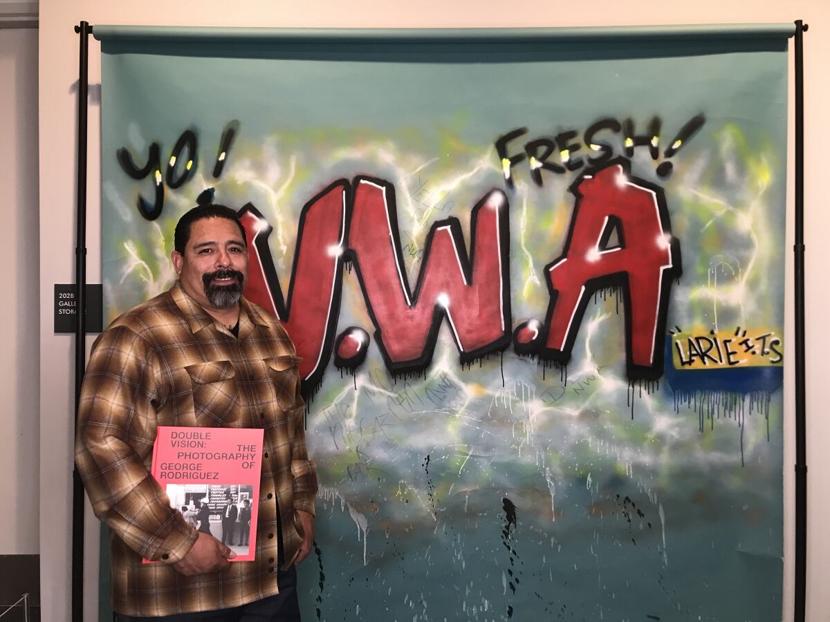 Larry Ruedas Jr. — once known as LARIE — stands in front of a graffiti backdrop he painted for N.W.A.