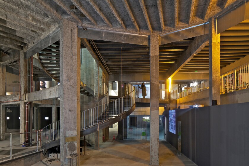 An interior view of the Palais de Tokyo shows an industrial, two-story space connected by a spiral stairway.