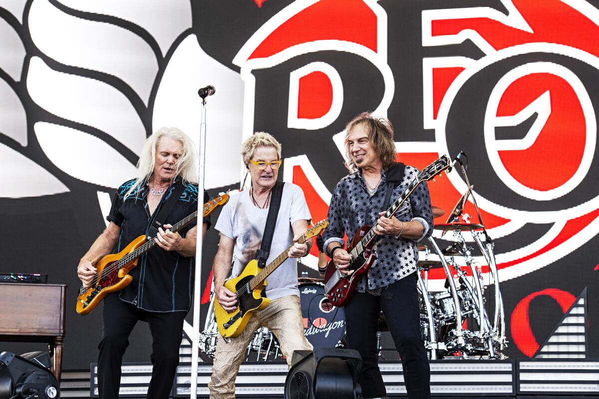 Three members of REO Speedwagon playing guitars together on an outdoor stage