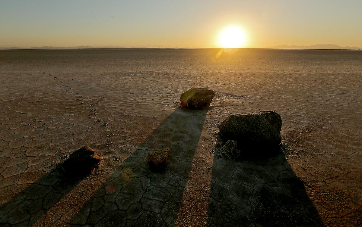 Boulders sit on an empty plain. In the background the sun sets on the horizon.