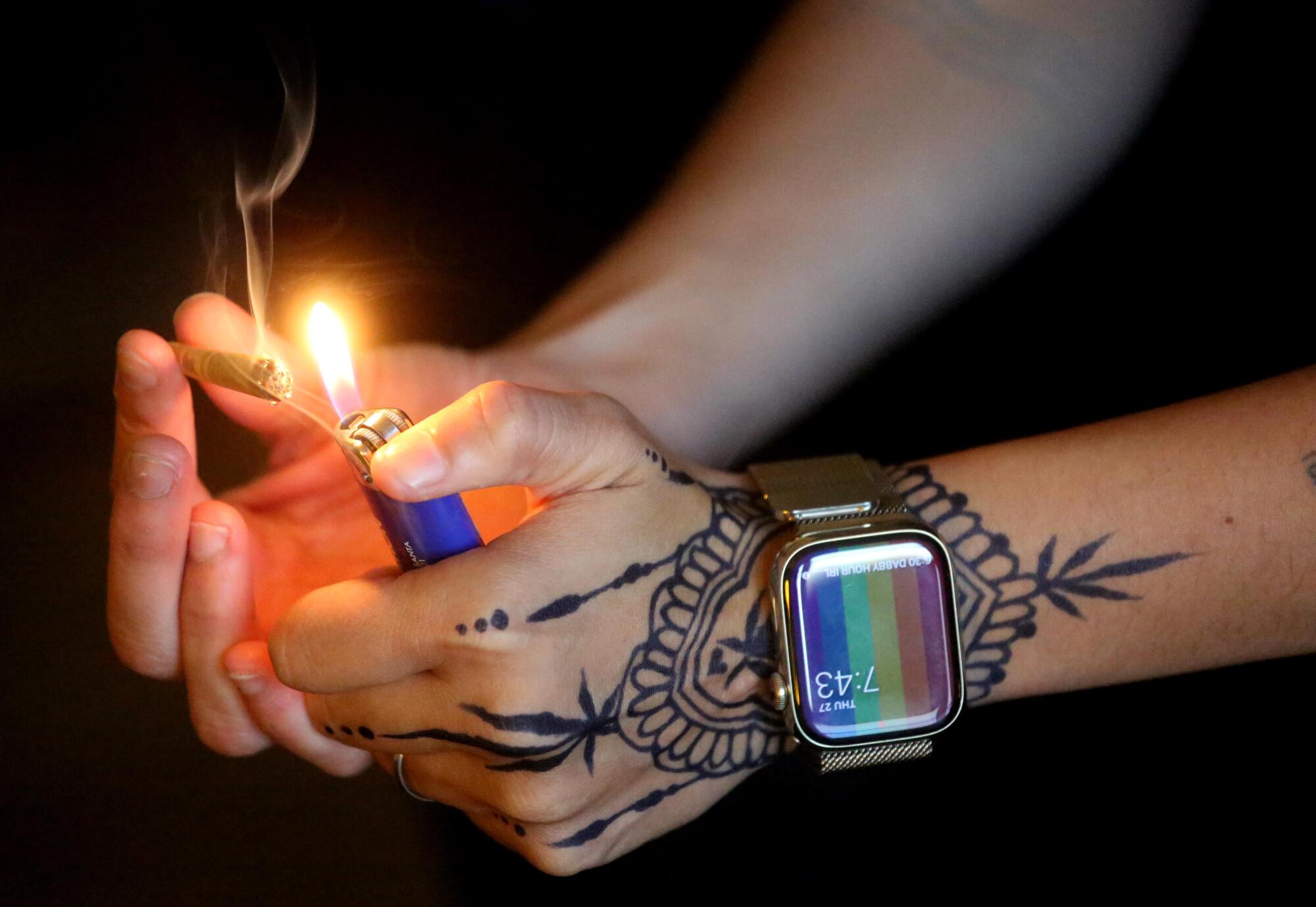 A closeup of two hands, one with henna tattoos and a large watch, lighting a joint with a plastic lighter