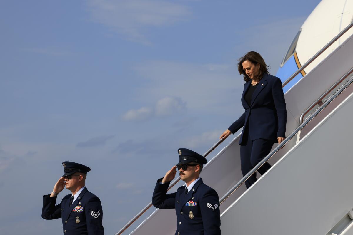 Kamala Harris walking down an airplane ramp as two men in navy-blue uniforms and hats stand below, saluting forward