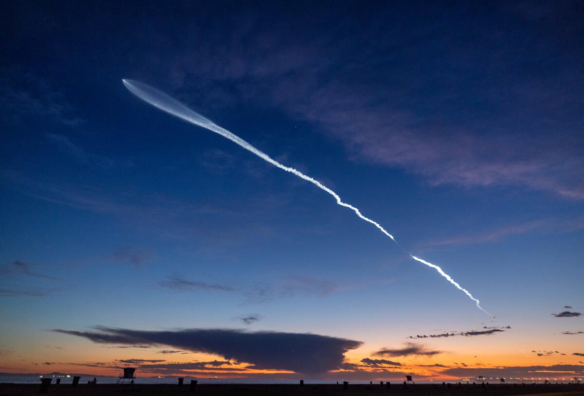 A contrail is seen across the sky at sunset along a coastline.