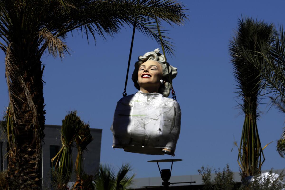 A portion of a statue of a woman hangs from moving equipment amid tall palms.
