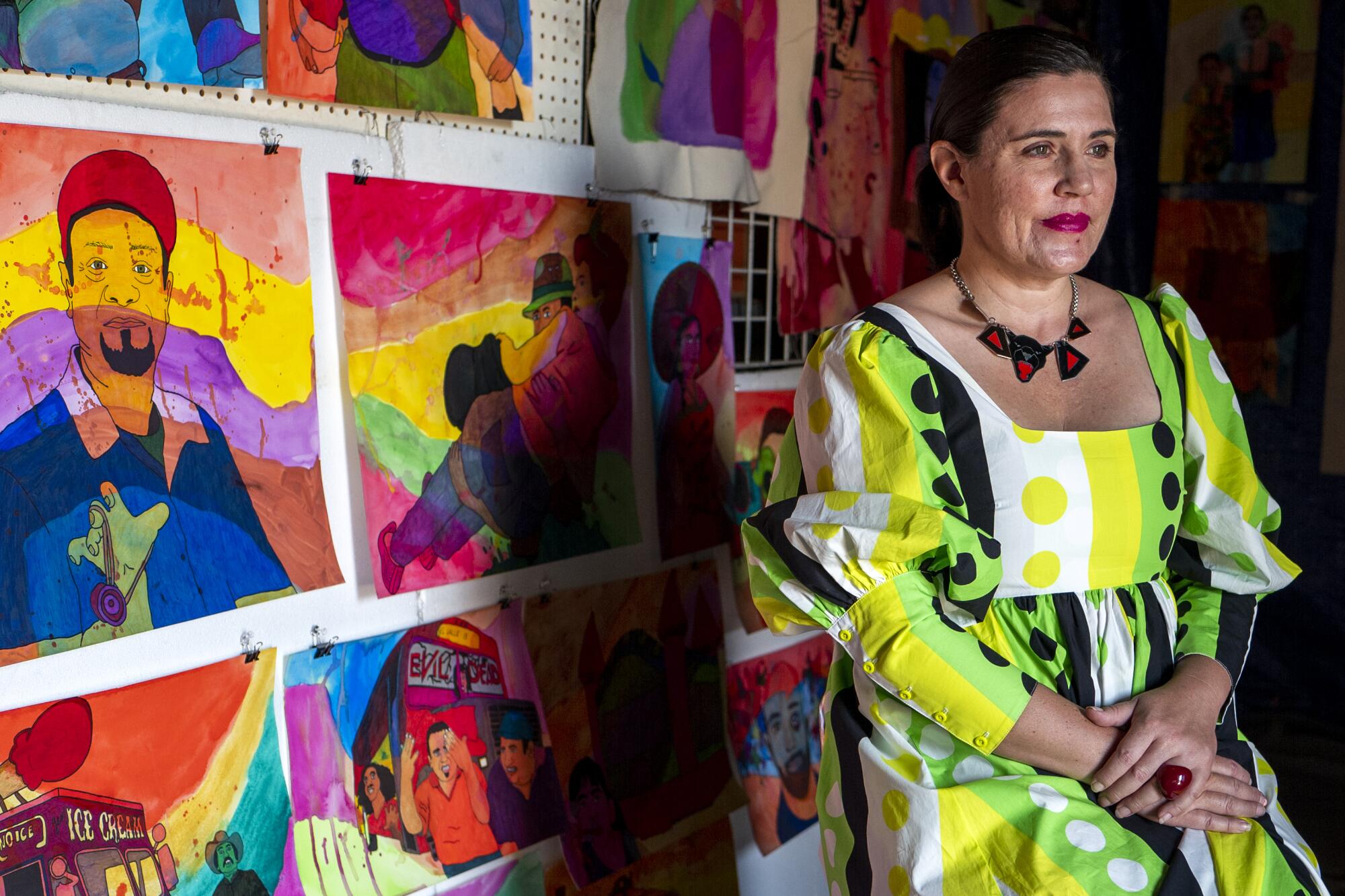 Karla Diaz, wearing a bright dress with a green and black geometric print, sits beside her watercolor paintings.