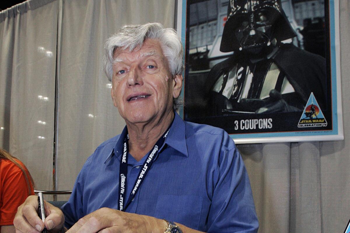 David Prowse sits with a poster of Darth Vader behind him.