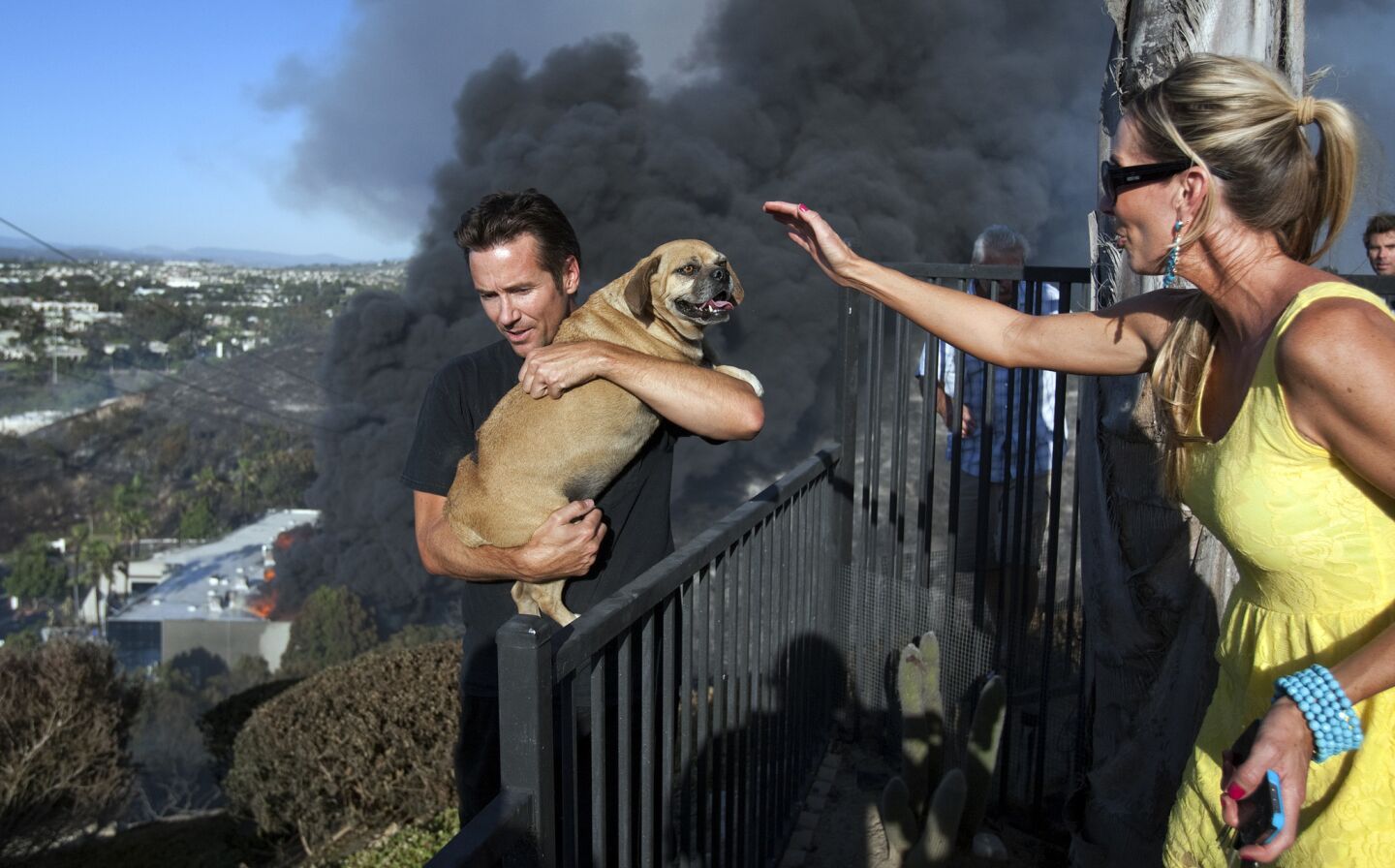Bronson Payne carries his dog, Rocky, to safety as his sister, Anya Bannasch, watches. Rocky was found hiding in the back of his burned home on Black Rail Road after it was destroyed in the Poinsettia wildfire on Wednesday in Carlsbad. An industrial blaze sparked by the wildfire burns in the background.