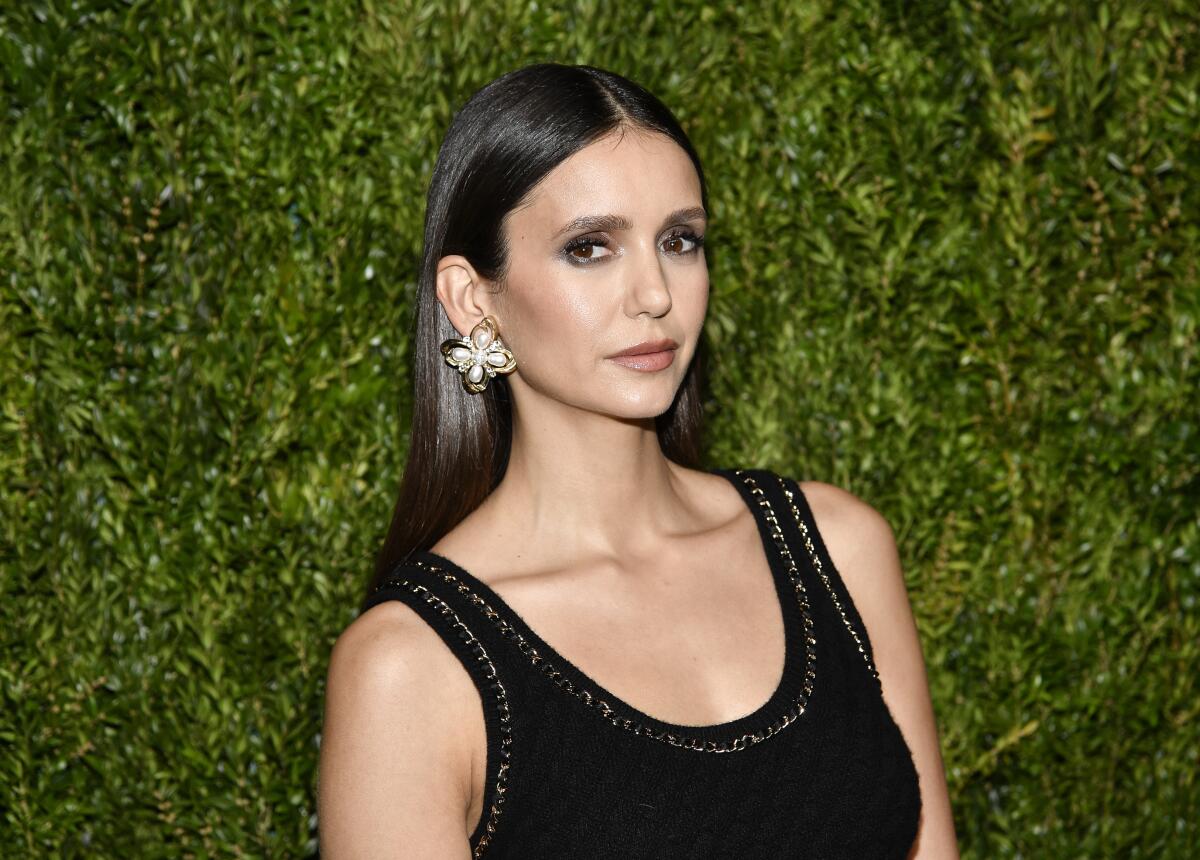 Nina Dobrev in black gown and floral earrings standing in front of a greenery wall