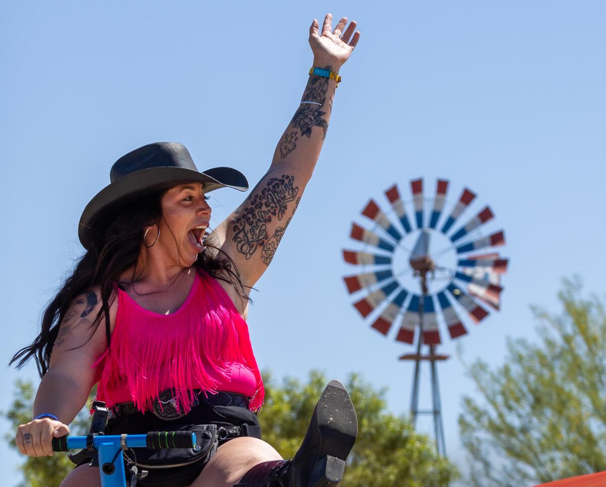 A woman wearing a cowboy hat waves her hand in the air atop a seesaw with a red, white and blue windmill in the background