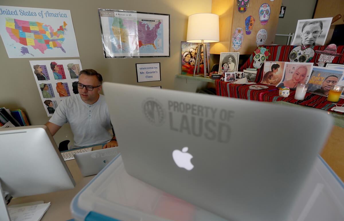 A teacher sits in front of multiple computer screens in his garage, where he's set up a small classroom space