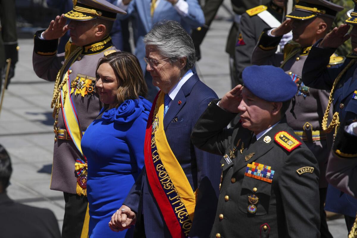 A gray-haired man hold hands with a woman in a blue dress as they walk escorted by two saluting men in military uniform.