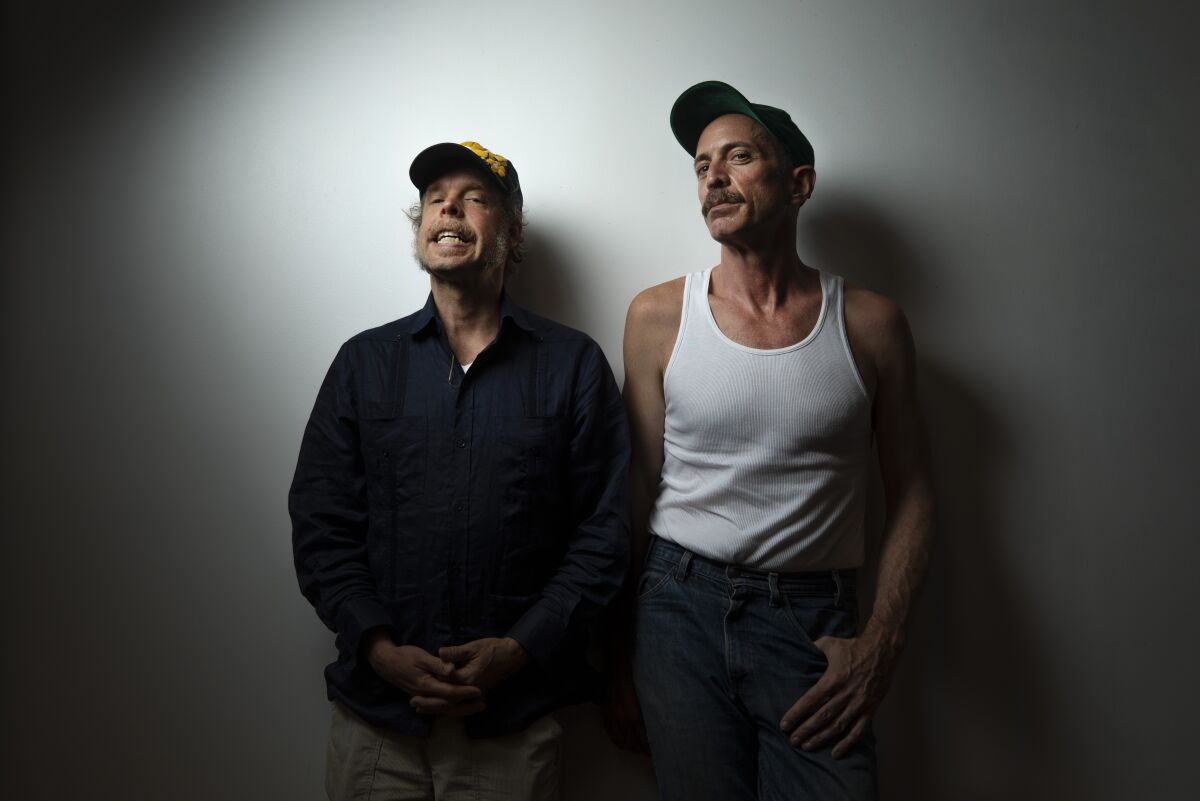 Two men stand, one with his fingers interlaced and another with a hand in his pocket