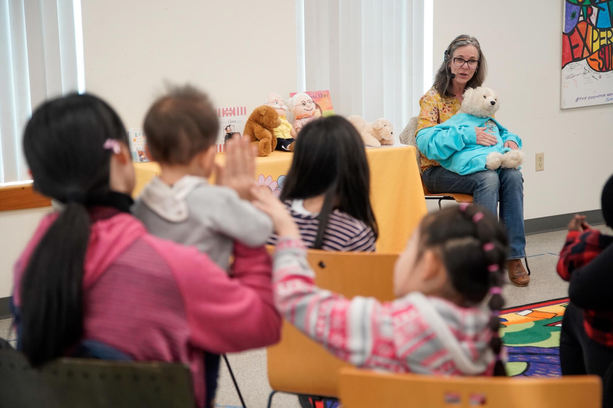 A woman holding a teddy bear sits in a chair speaking to a room full of seated toddlers and children.