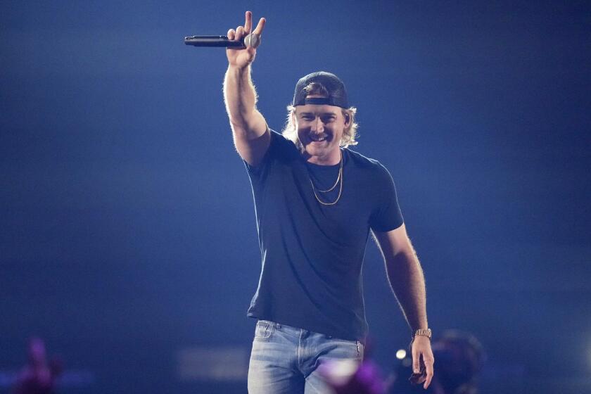 Morgan Wallen holds a microphone and waves to a crowd while wearing a black T-shirt, backward hat and jeans.