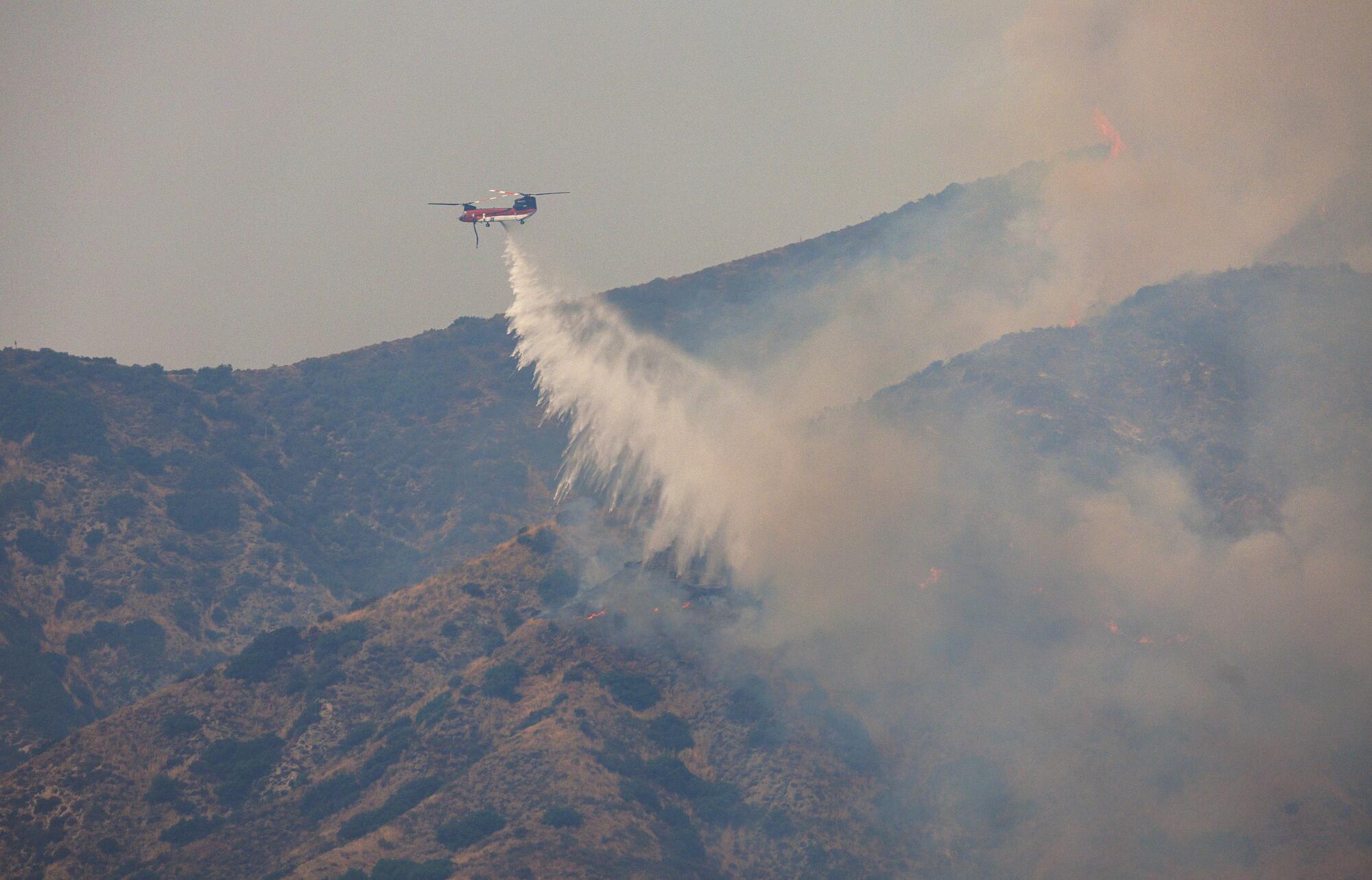 A helicopter drops water across a smoky hillside