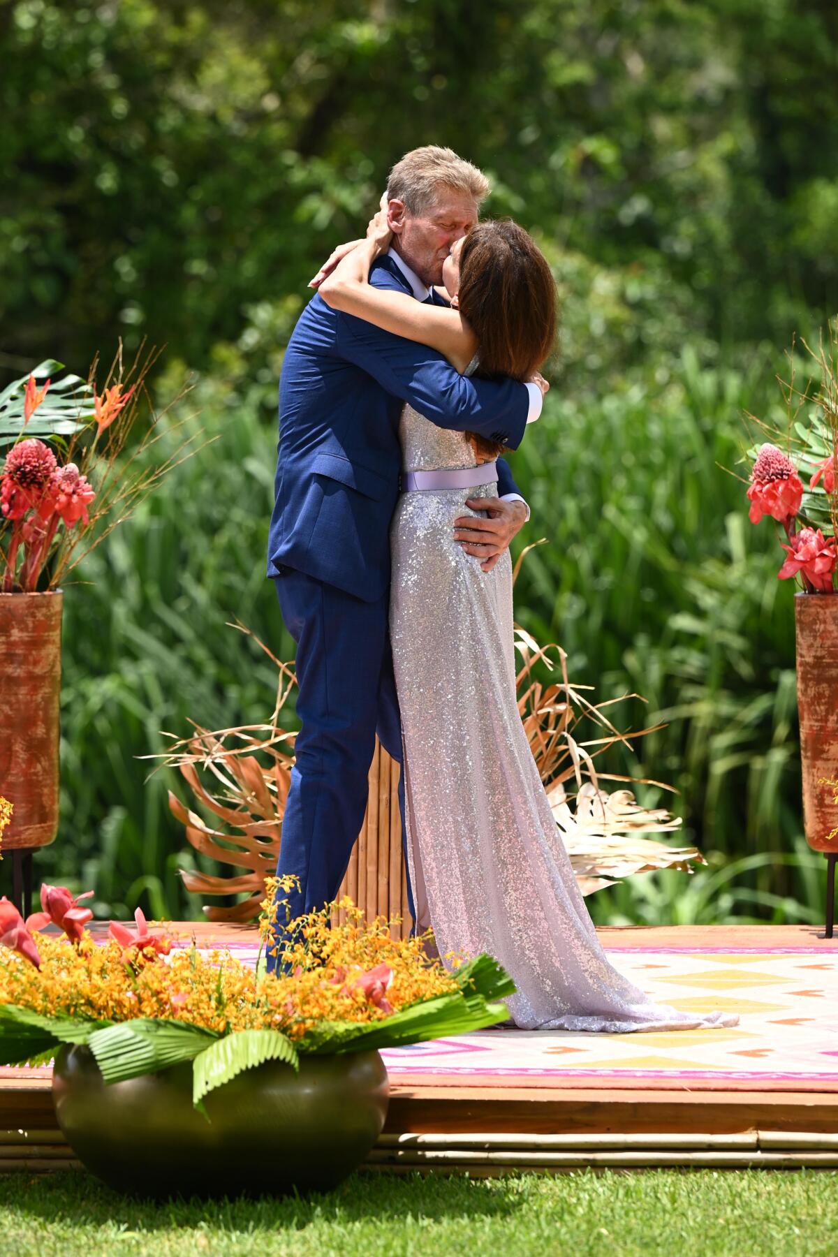 Standing outdoors on a platform, Gerry in a blue suit hugs Theresa in a silver dress.