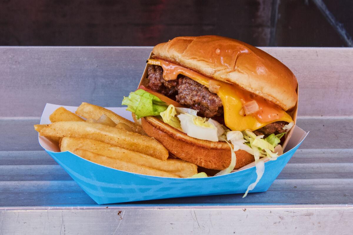 A fully dressed cheeseburger sits atop an order of fries in a blue paper basket.