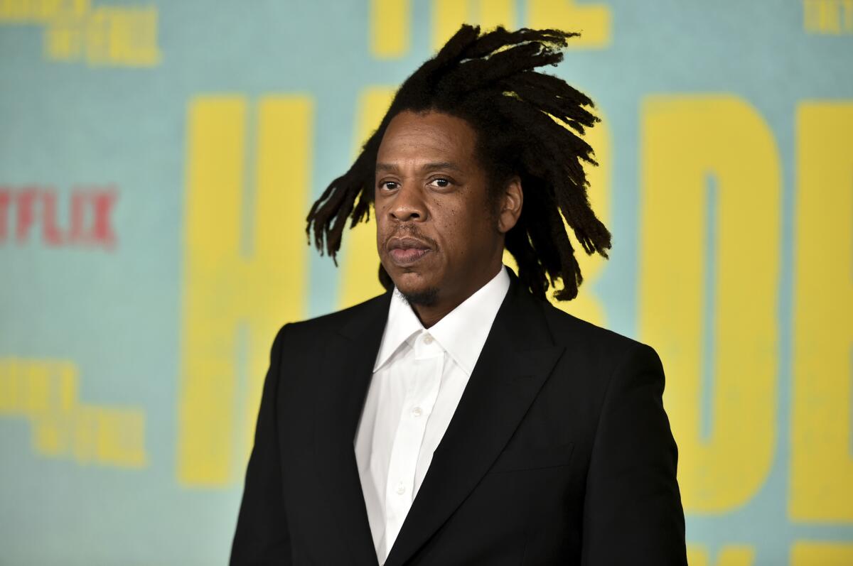 Jay-Z wears a black suit jacket and white button-up dress shirt with his hair in free-form dreadlocks