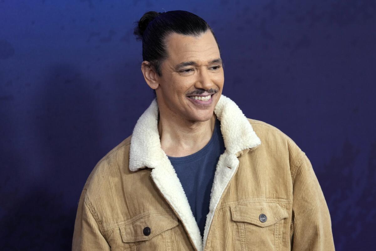 A man with black hair pulled up in a bun and a mustache smiles in a beige jacket and blue shirt