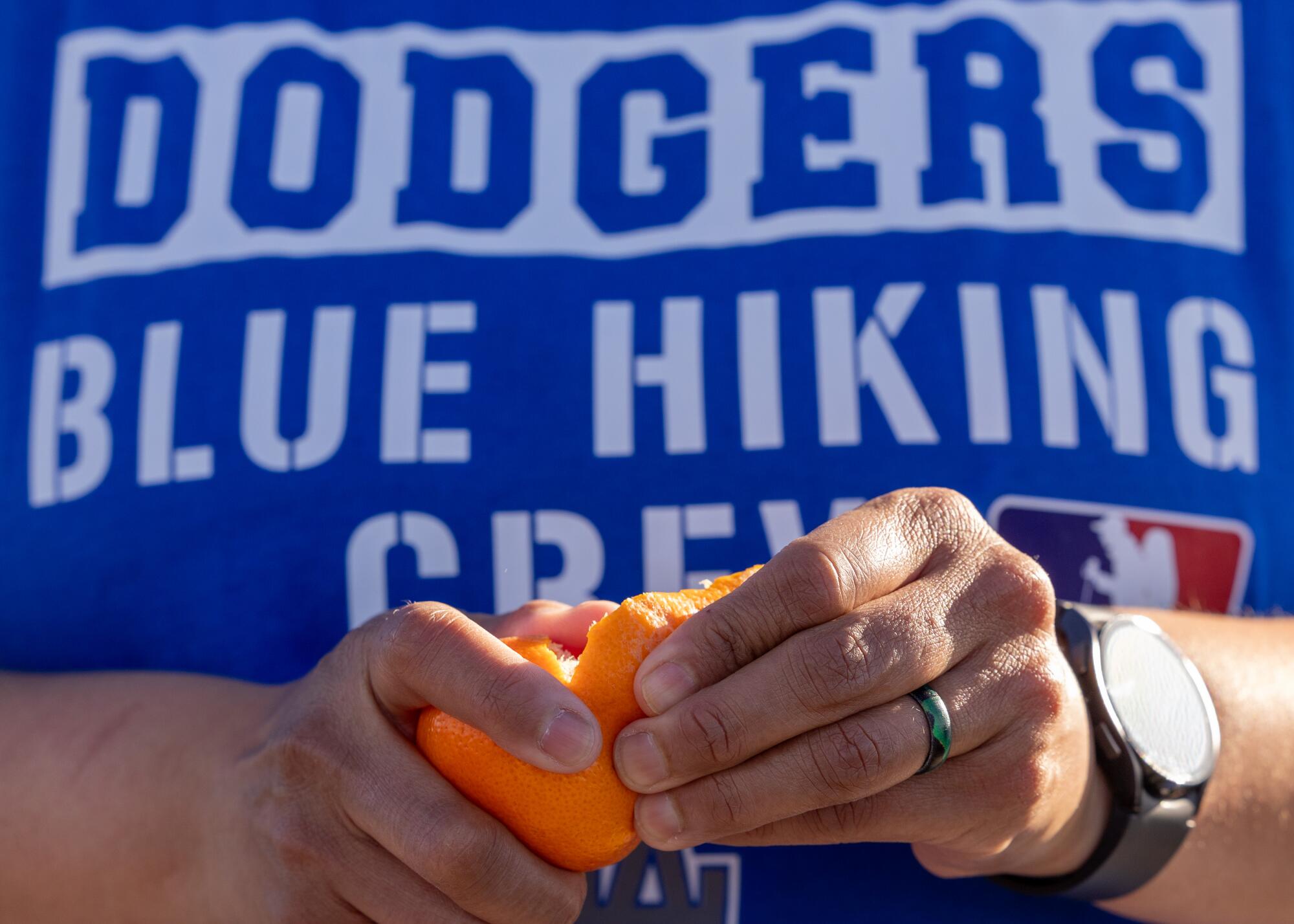A hiker wearing custom Dodgers Blue Hiking Crew merchandise takes a break to peel a tangerine sandwich.