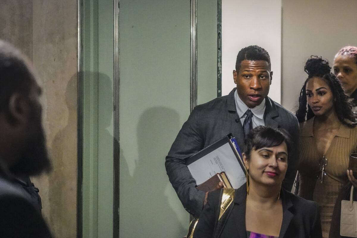 Jonathan Majors in a grey suit next to a glass door entering a court room surrounded people 
