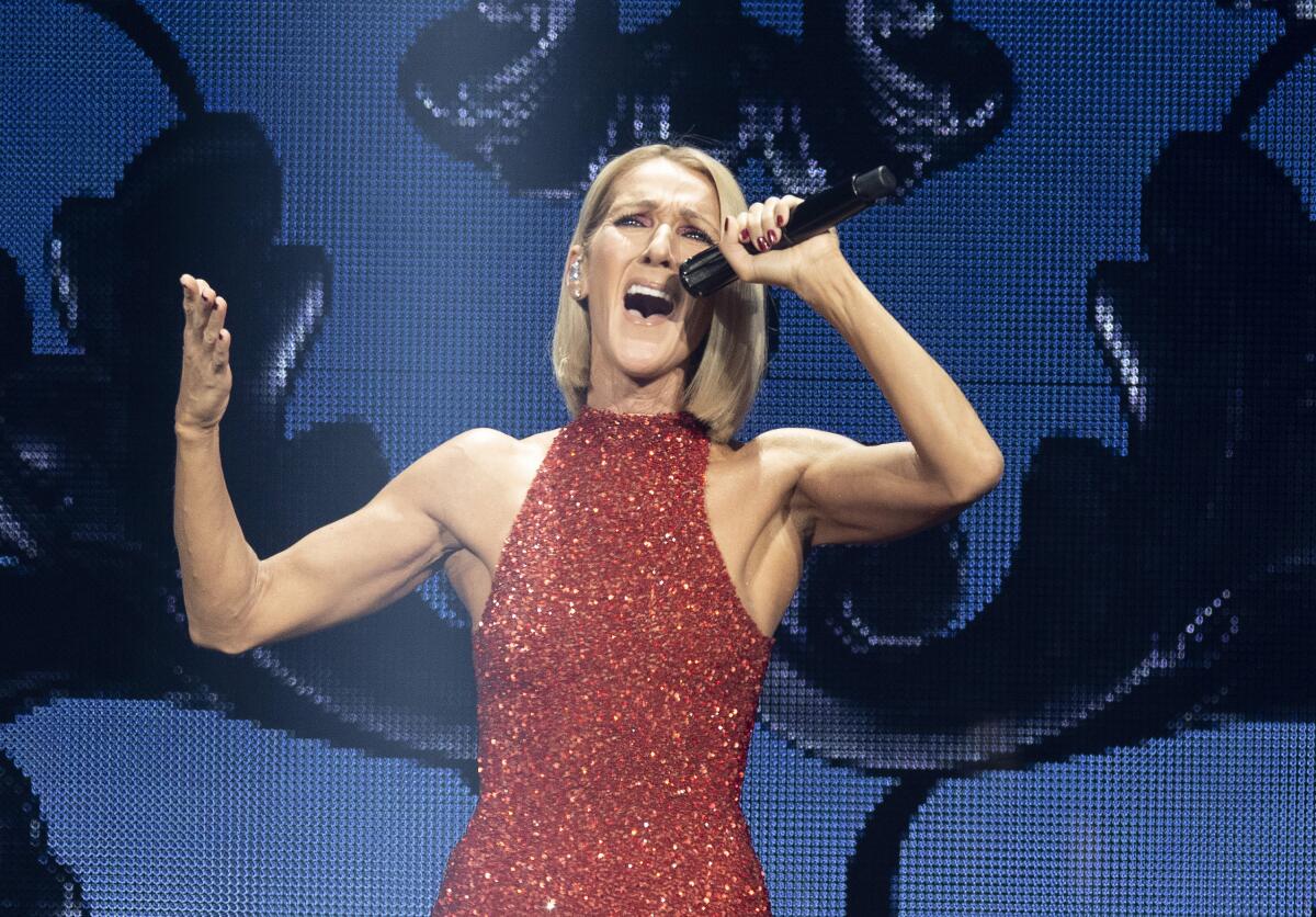 Celine Dion wearing a sparkling red gown sings onstage with her mouth wide open and her arms raised