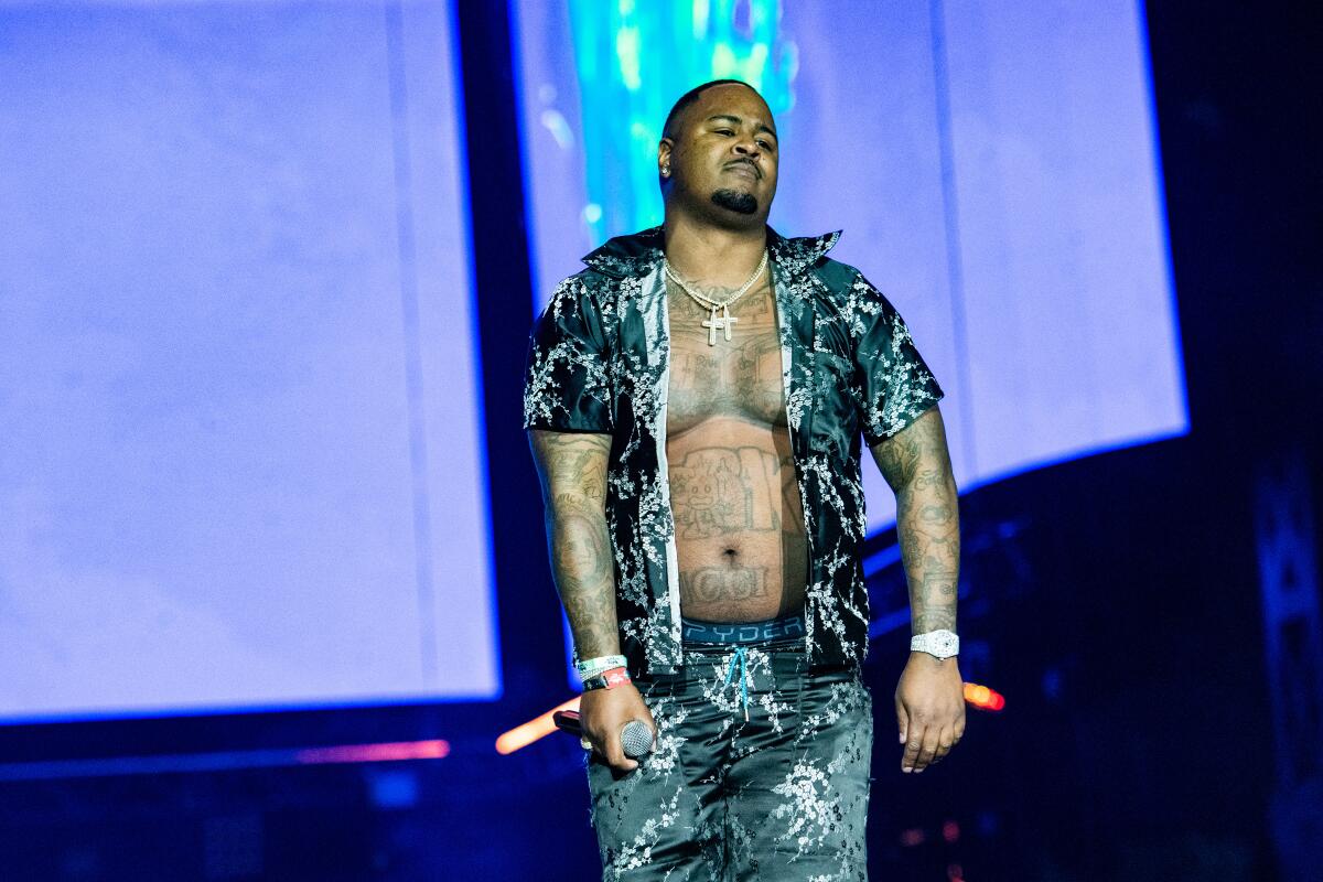 Drakeo the Ruler, unbuttoned shirt revealing tattoos, stands onstage with microphone in hand