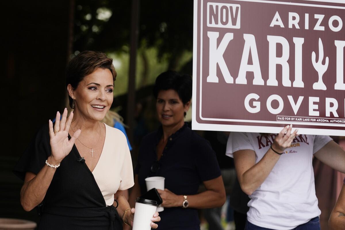 Kari Lake waves to supporters as someone beside her holds up a large campaign sign. 