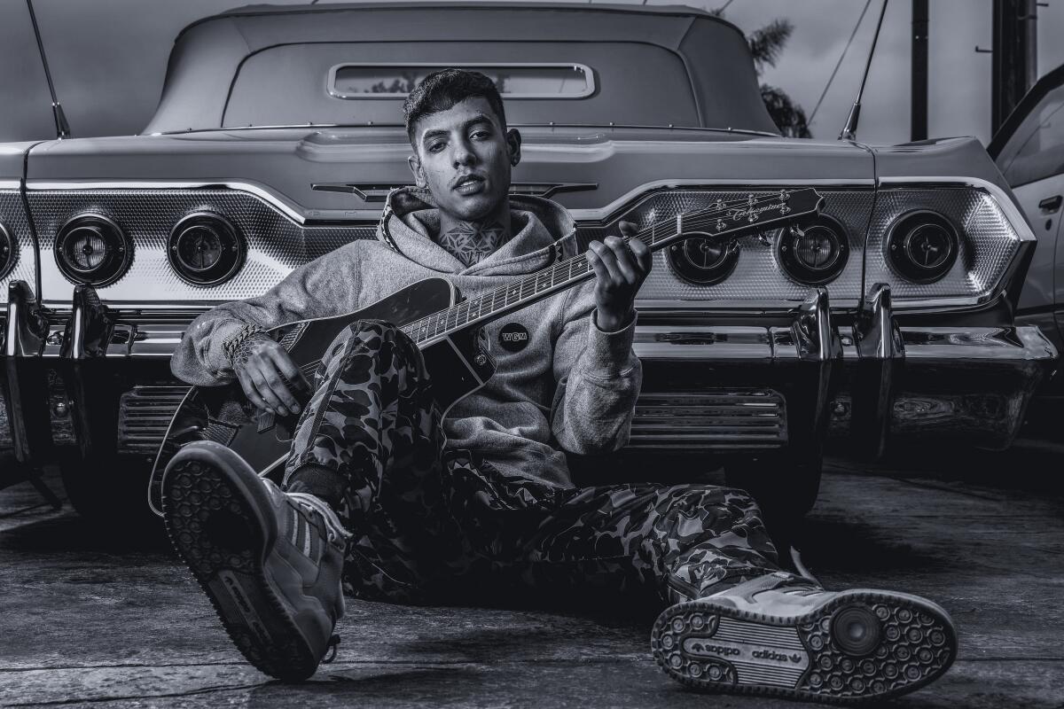 A man sits with a guitar, his back on a car
