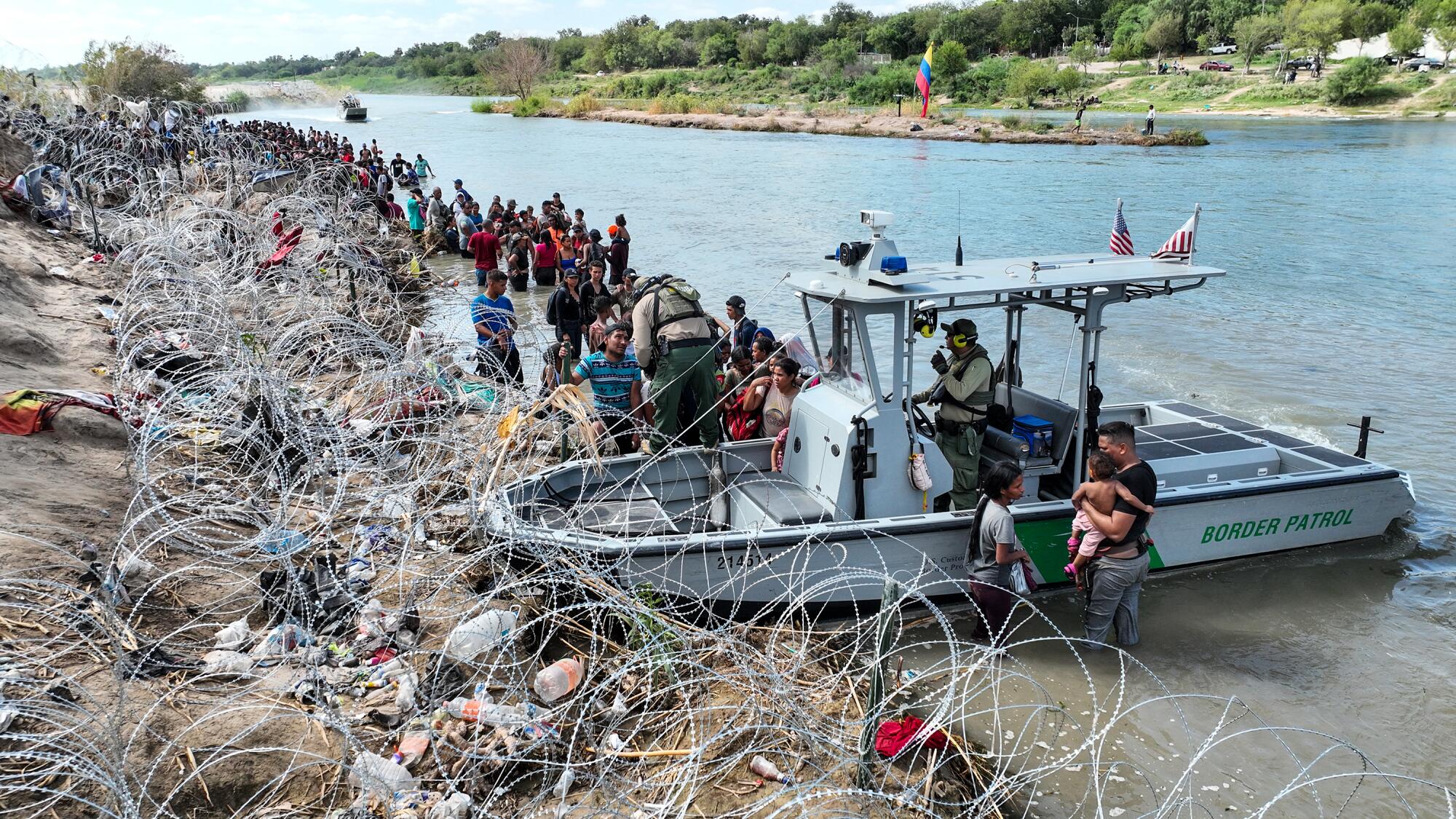 A Border Patrol boat sits next to coils of razor wire as a long line of people stand in the river.