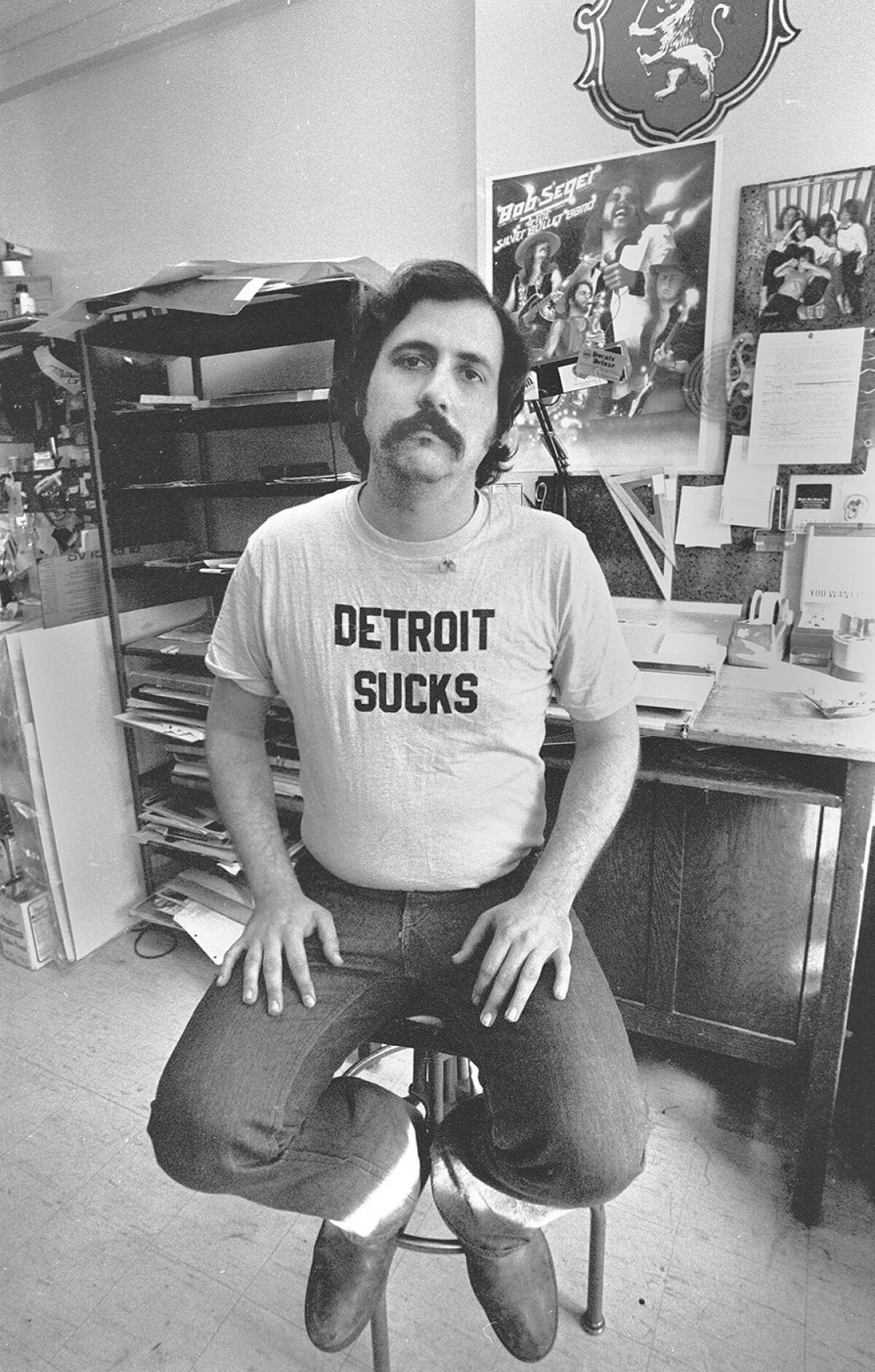 A black-and-white photo of a mustached man in a T-shirt that says "Detroit Sucks," sitting on a stool.