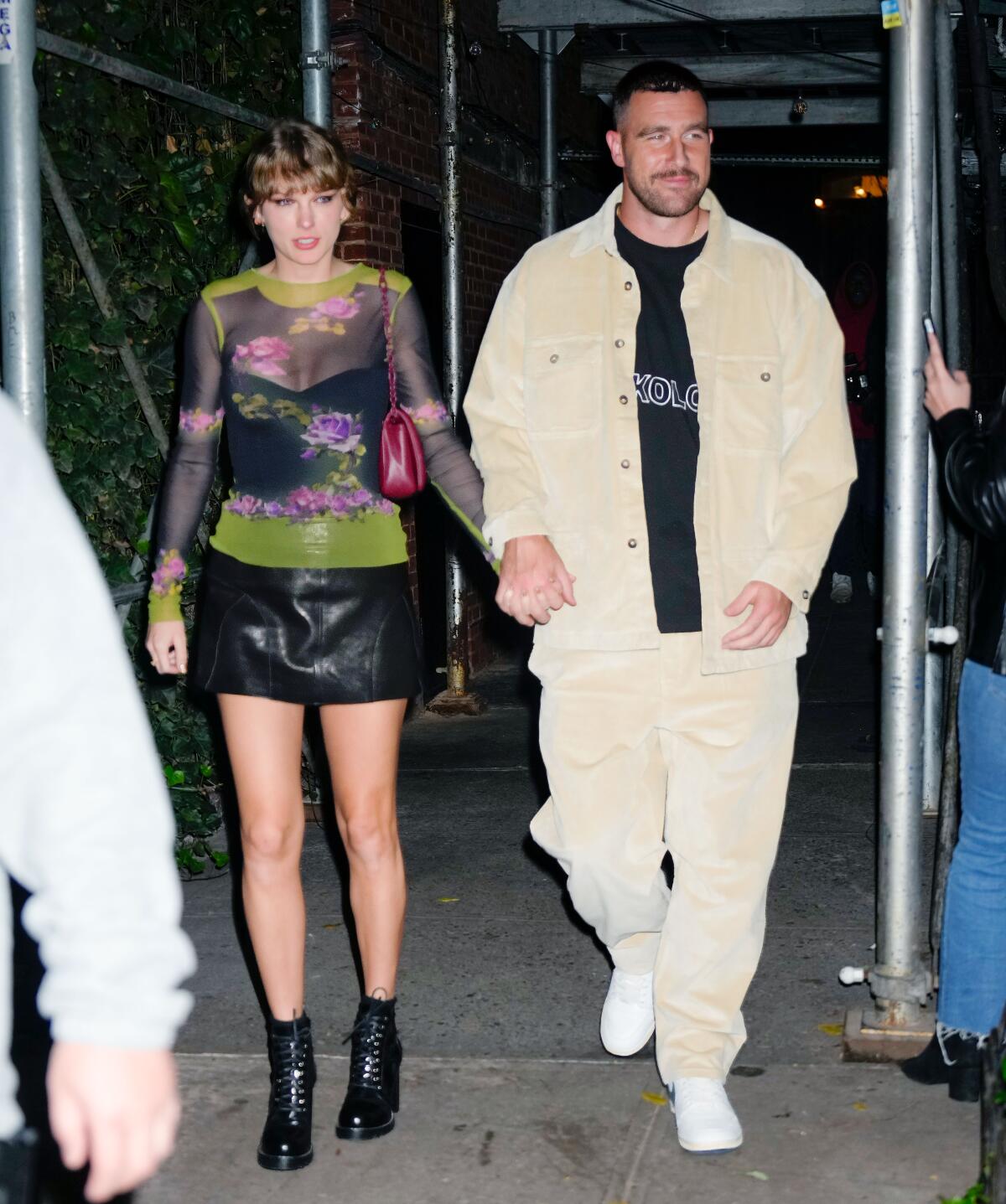  Taylor Swift and Travis Kelce hold hands while walking on a sidewalk under scaffolding