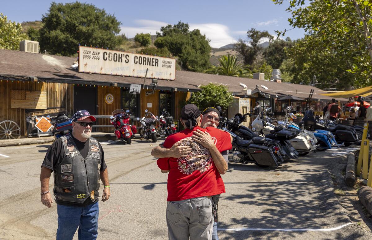 Two men hug as a third looks on in a parking lot filled with motorcycles