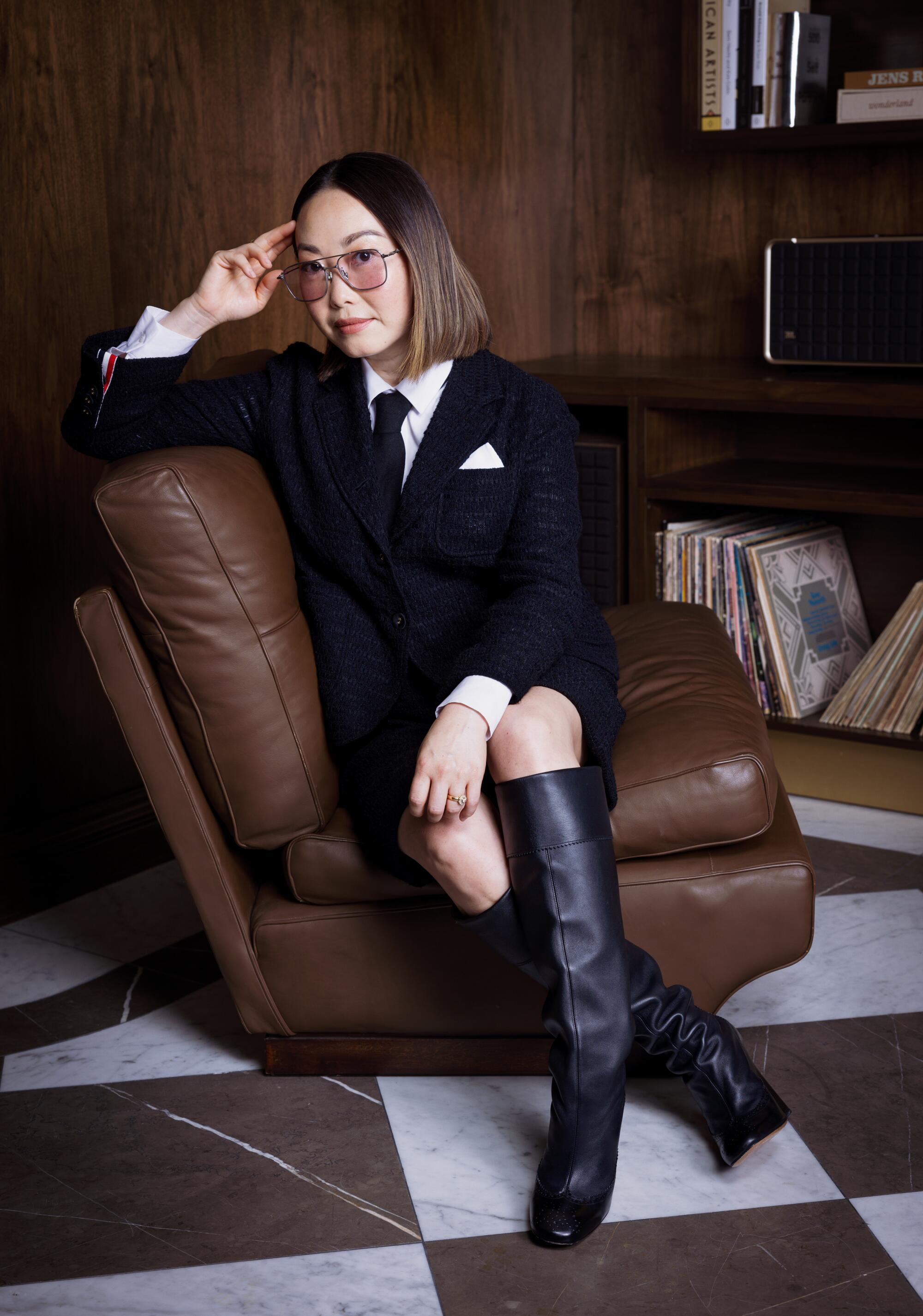 Director Lulu Wang, in a skirt suit and tall boots, sits sideways in a leather chair for a portrait.