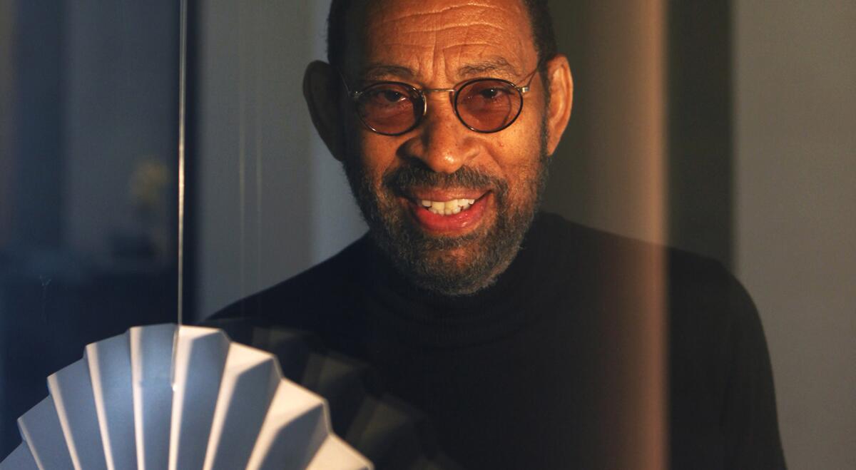 Maurice Hines Jr. sits in a black sweater and round tinted glasses with a white fan in the foreground