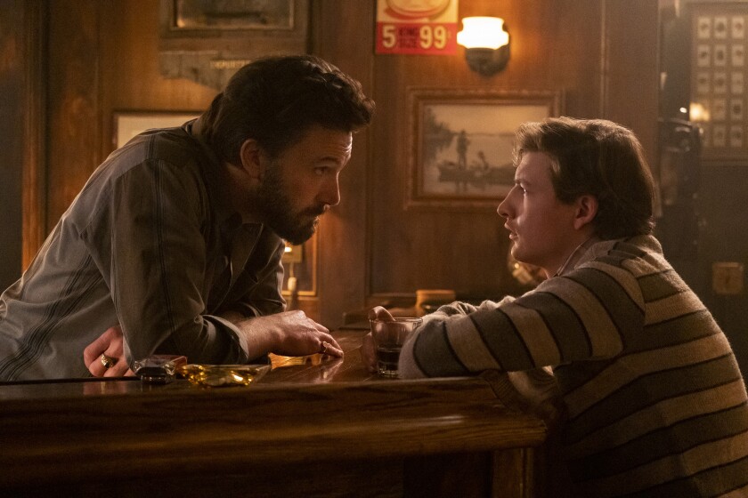 An older and a younger man talk to each other across a bar.