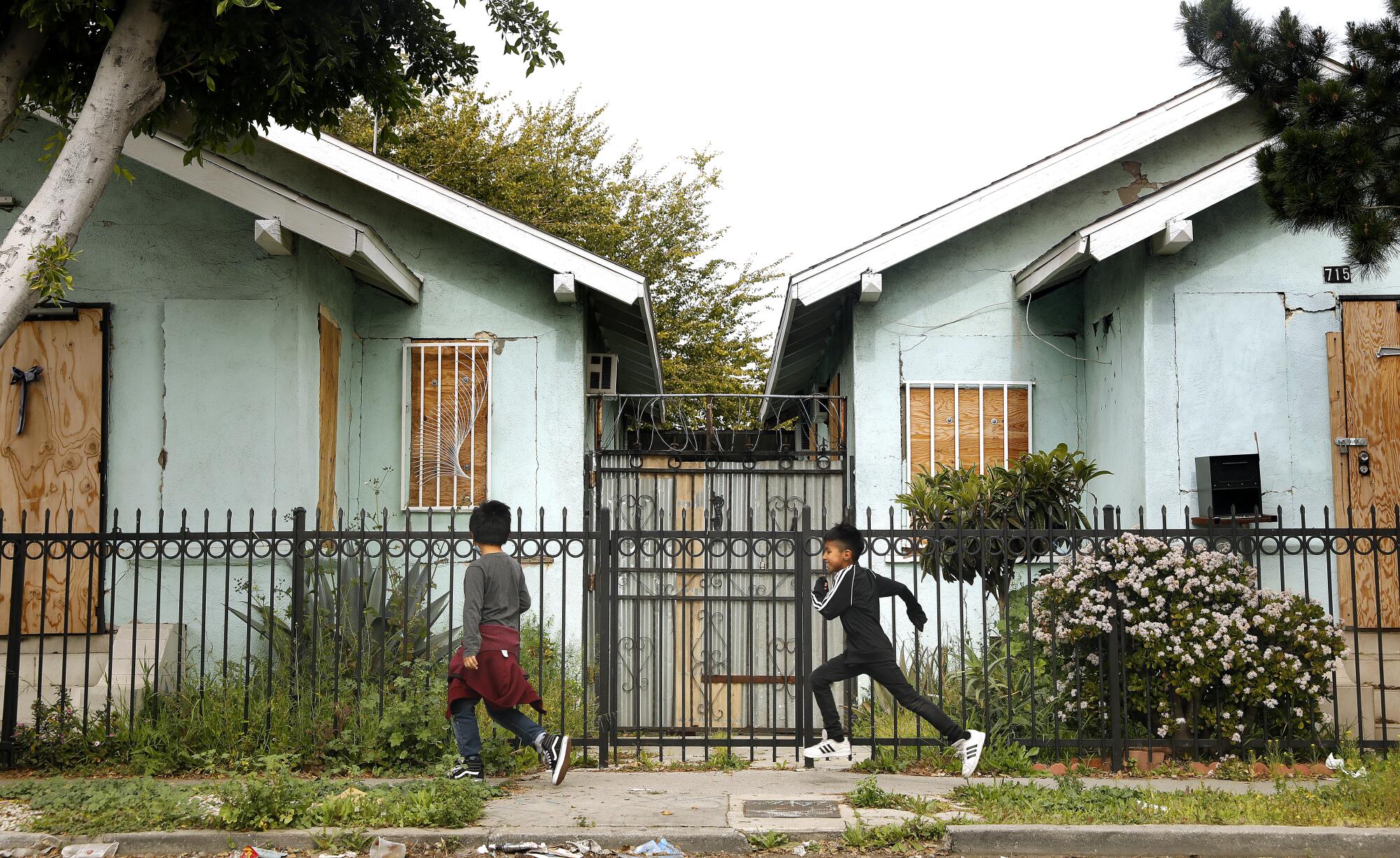 Two kids run past a metal fence and boarded up homes 