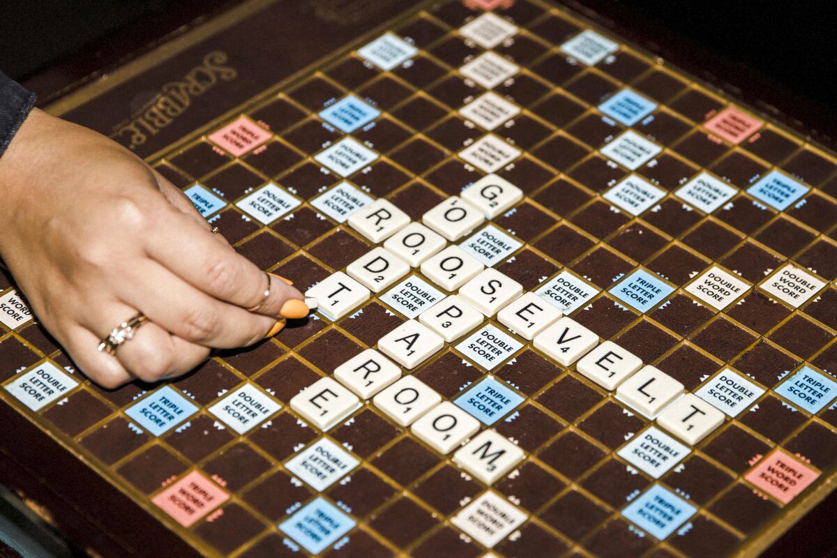 A hand with a diamond ring and yellow nail polish puts down letters in a game of Scrabble.